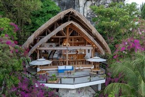 Una cabaña de madera con techo de paja, mobiliario exterior y rodeada de exuberante vegetación.