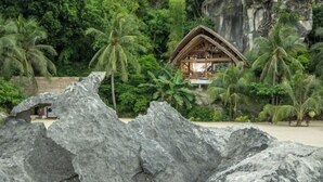 Een houten huis met rieten dak, omgeven door weelderige begroeiing en een rotsachtige voorgrond.
