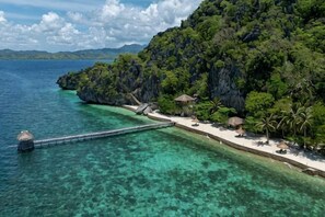 Une zone côtière avec une jetée s’avançant dans une eau claire, entourée d’une végétation luxuriante et bordée d’une petite plage où se trouvent des cabanes aux toits de chaume.
