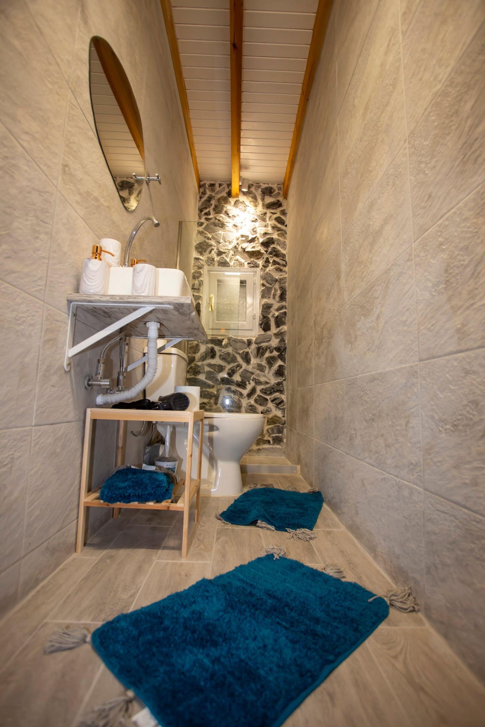 A narrow bathroom with a stone wall, a wooden shelf with toiletries, and a blue rug on the floor.