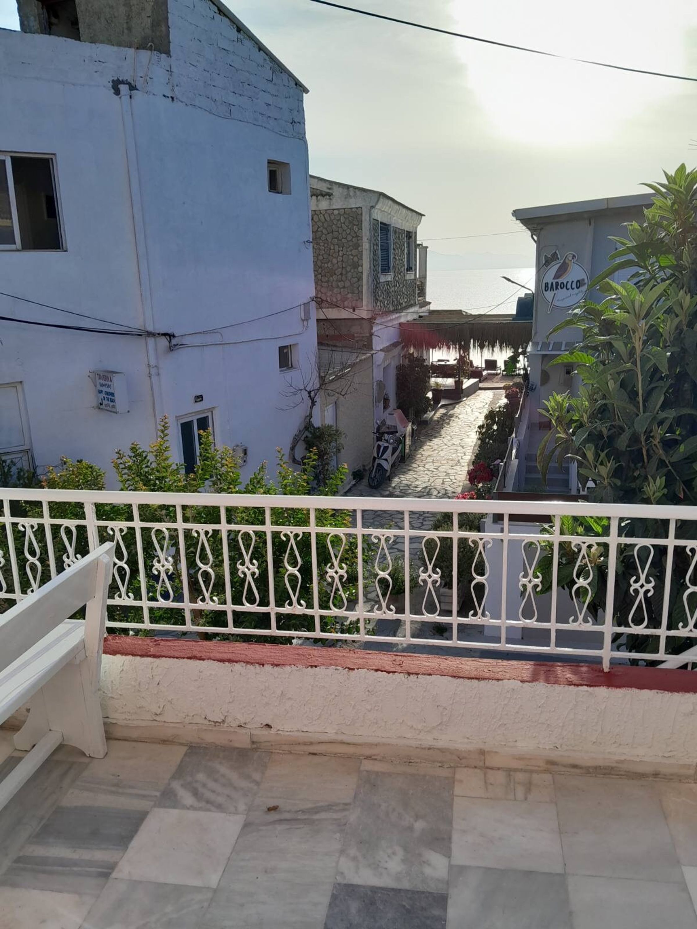 A balcony with a tiled floor, a white railing, and a view of a narrow street with buildings and a parked scooter.
