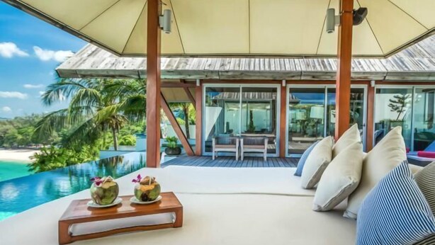 A luxurious outdoor seating area with a wooden table, cushioned seating, and a view of a tropical beach and ocean. - Laemsingh Villa 3 (Phuket)