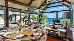 A dining area with round tables set for a meal, large windows offering a view of the sea, and a ceiling with wooden beams. - Laemsingh Villa 3 (Phuket)