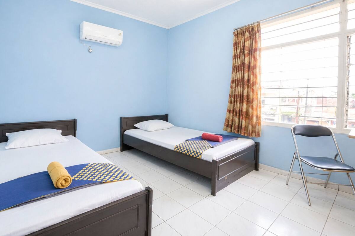 Una habitación con dos camas, una silla, un ventanal con cortinas y un equipo de aire acondicionado.