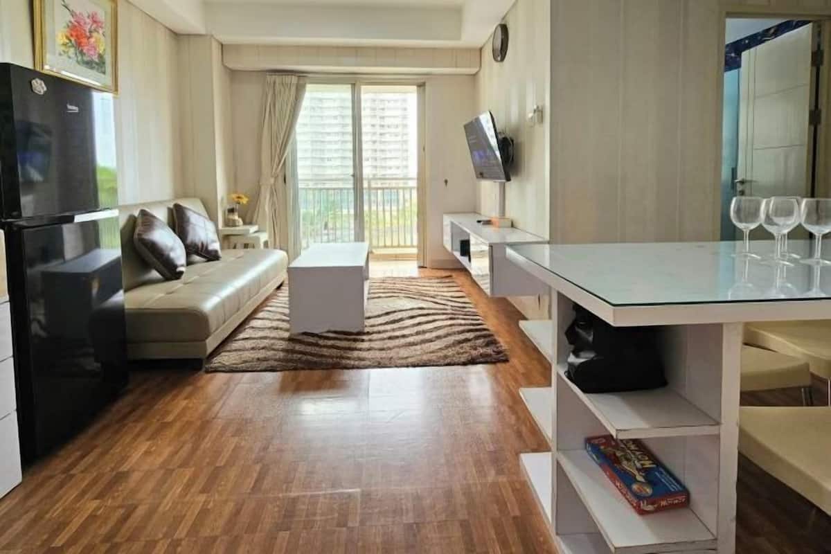 A modern living room with a sofa, a flat-screen TV, a dining table with chairs, and a kitchen area with a refrigerator.