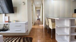 A hotel room with a flat-screen TV, a desk with chairs, a white wardrobe, and a built-in shelf unit. - Trivium Terrace Apartment Lippo Cikarang (Min Stay 3 Nights) (South Cikarang)