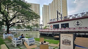 A riverside seating area with tables and chairs, a large boat with a logo, and a sign indicating boat service hours. - Trivium Terrace Apartment Lippo Cikarang (Min Stay 3 Nights) (South Cikarang)
