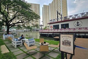 A riverside seating area with tables and chairs, a large boat with a logo, and a sign indicating boat service hours. - Trivium Terrace Apartment Lippo Cikarang (Min Stay 3 Nights) (South Cikarang)