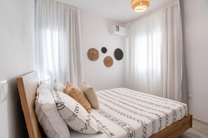 Ein Bett mit einem gemusterten Überwurf, weißen Kissen und einem hölzernen Kopfteil. An der Wand befinden sich zwei Klimaanlagen und ein gewebter Wandbehang über dem Bett.
