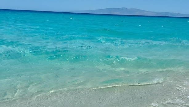 Vista despejada de una playa con aguas turquesas y una cadena montañosa distante.