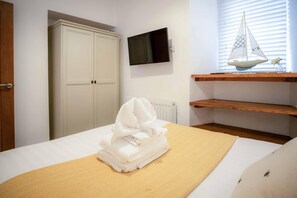A bedroom with a bed, a white wardrobe, a TV mounted on the wall, and a window with blinds. - Caldey View - 3 Bed - Hot Tub - Penally - Tenby (Tenby)