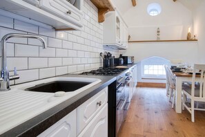 Una cocina con armarios blancos, encimera negra y suelo de madera. Hay una ventana sobre el fregadero y una zona de comedor con sillas blancas.