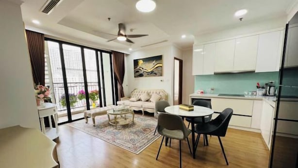 A modern living room with a sofa, coffee table, and dining area. There is a large window with curtains, a ceiling fan, and a kitchen area with cabinets and appliances. - Vinhomes Royal City Apartments - Sunhometel (Hanoi)
