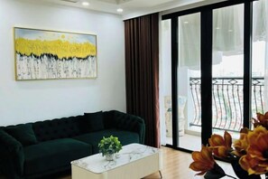 A living room with a dark green sofa, a white coffee table with a plant, and a large window with a view of a balcony.