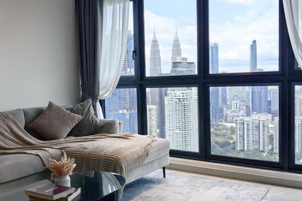 Ein modernes Wohnzimmer mit großem Fenster, einem grauen Sofa und Blick auf hohe Gebäude.