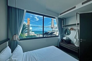 Une chambre d’hôtel comprenant un lit, une armoire, un miroir, une fenêtre donnant sur l’océan et les bâtiments, ainsi qu’un luminaire suspendu.