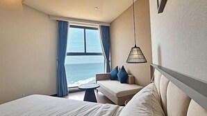 Une chambre d’hôtel avec un lit, un canapé avec des coussins bleus, une petite table ronde et une vue sur l’océan par une grande fenêtre ornée de rideaux bleus.