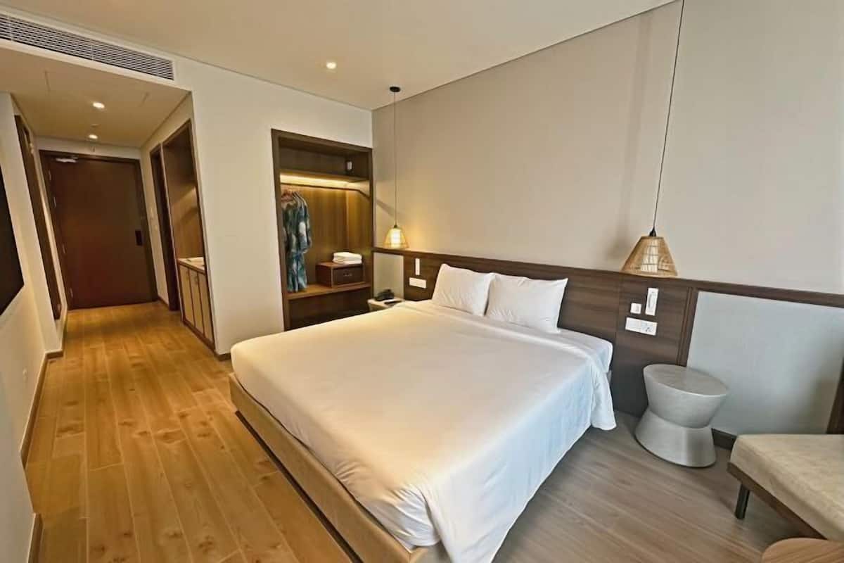 A hotel room with a large bed, a bedside table, a chair, a wardrobe, and a small stool.