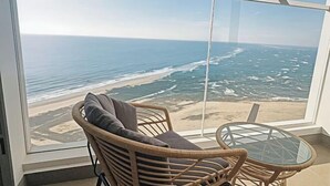 Un balcon avec vue sur l’océan et la plage, des meubles en osier et une table en verre.