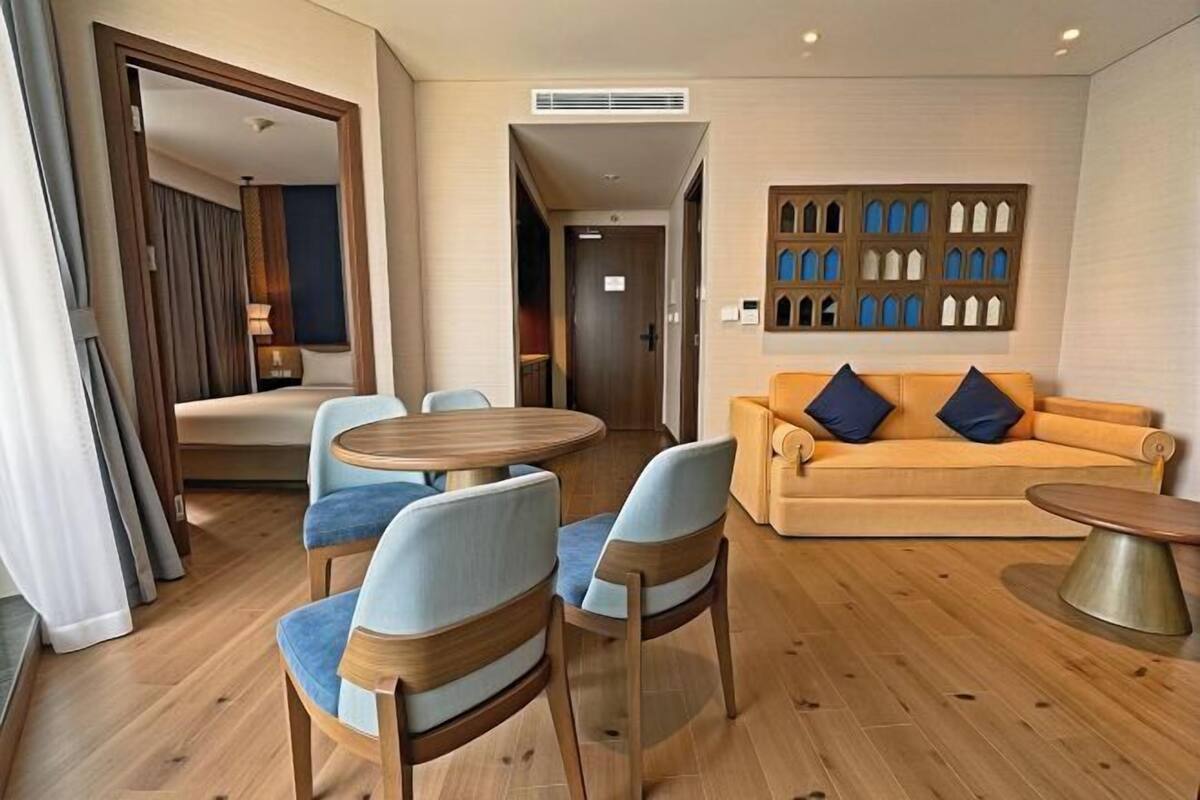 A hotel room with a wooden floor, a round dining table with chairs, a sofa with cushions, and a wall with a decorative panel.