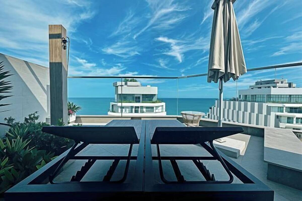 A rooftop terrace with a table and chairs, a wooden column, and a view of the ocean and buildings.