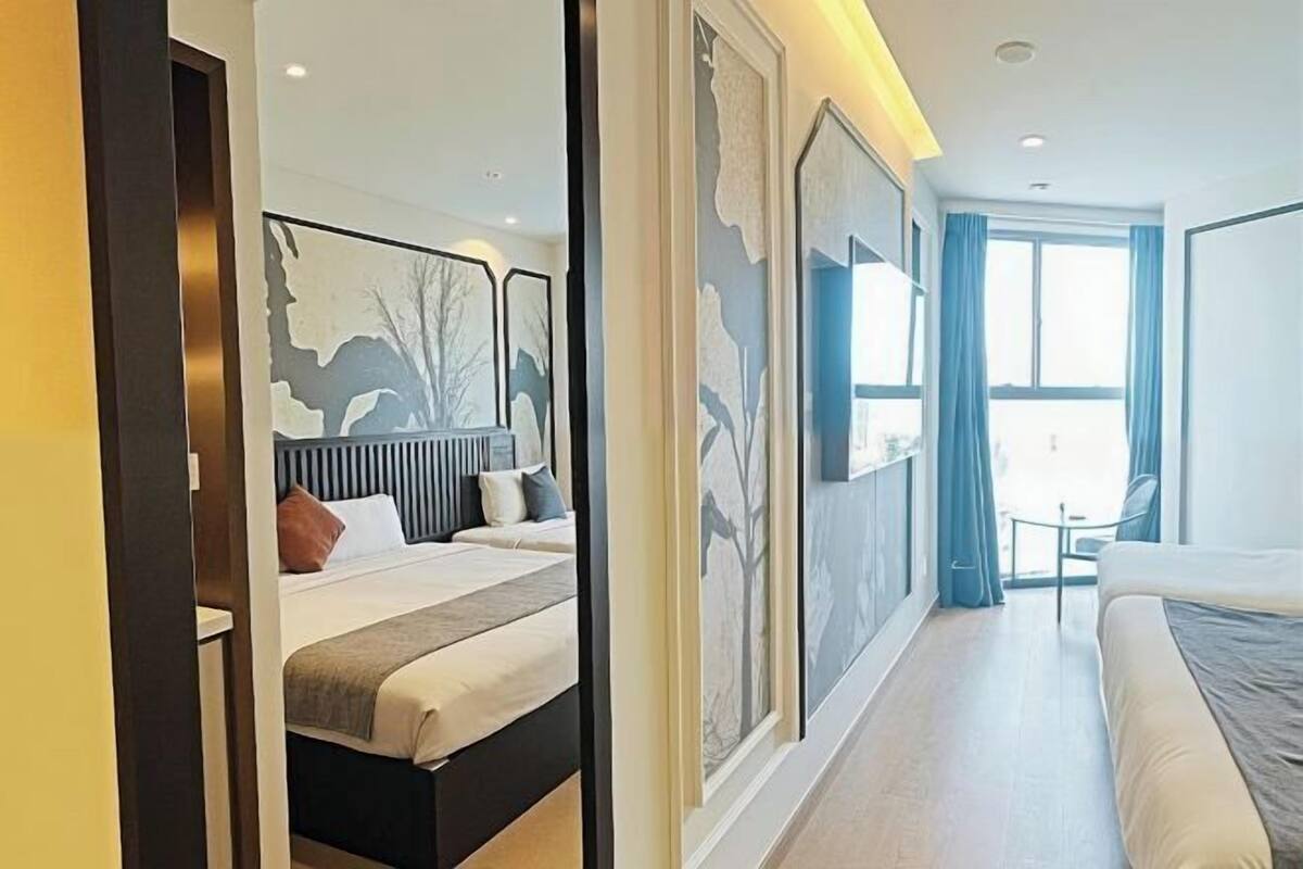A hotel room with a large bed, a smaller bed, a desk, and a chair. There are large windows with curtains.