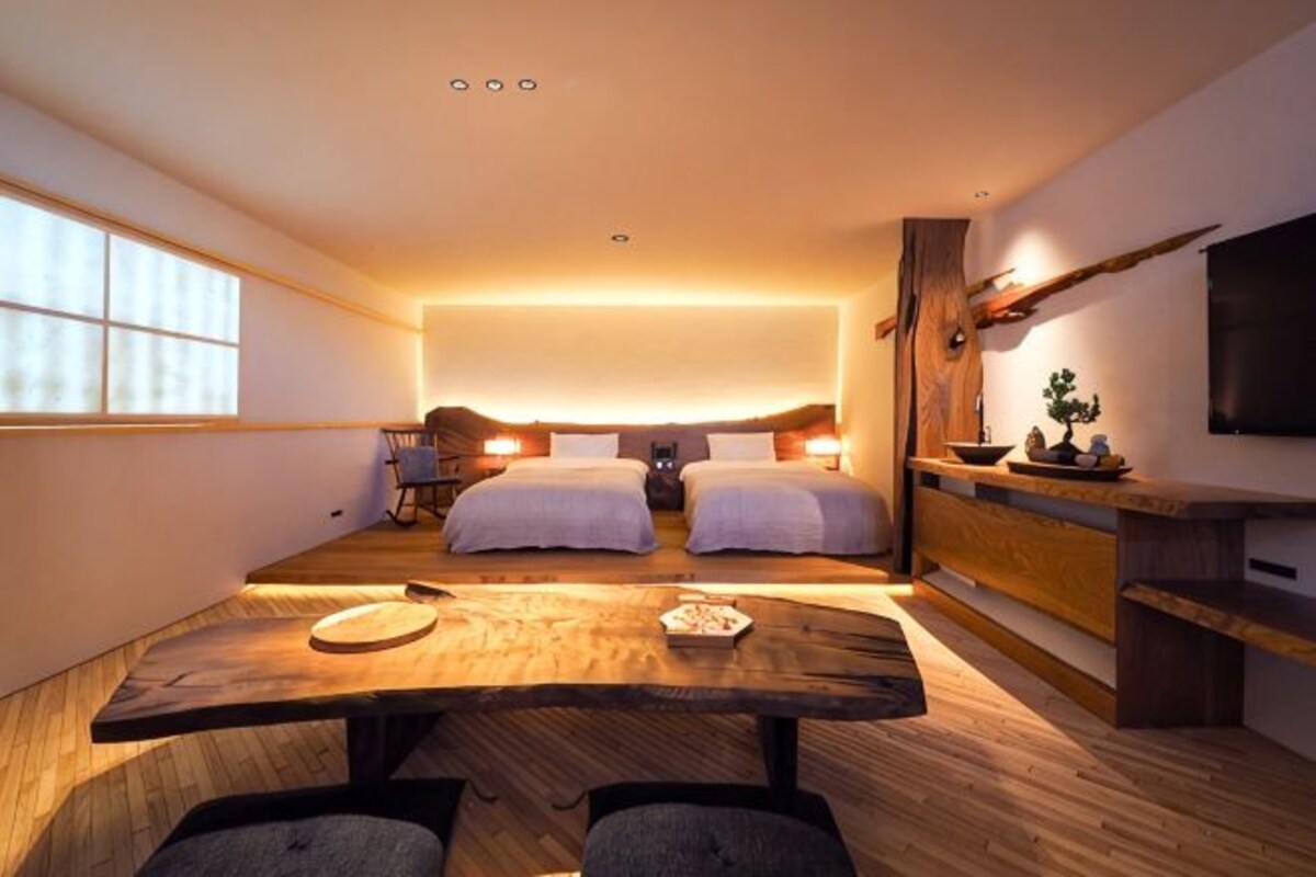 A compact room with two beds, a dining table, a TV, and a small plant.