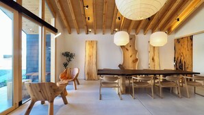 Et moderne spiseområde med et træbord og stole, store vinduer samt et loft med synlige bjælker og hængende lamper.