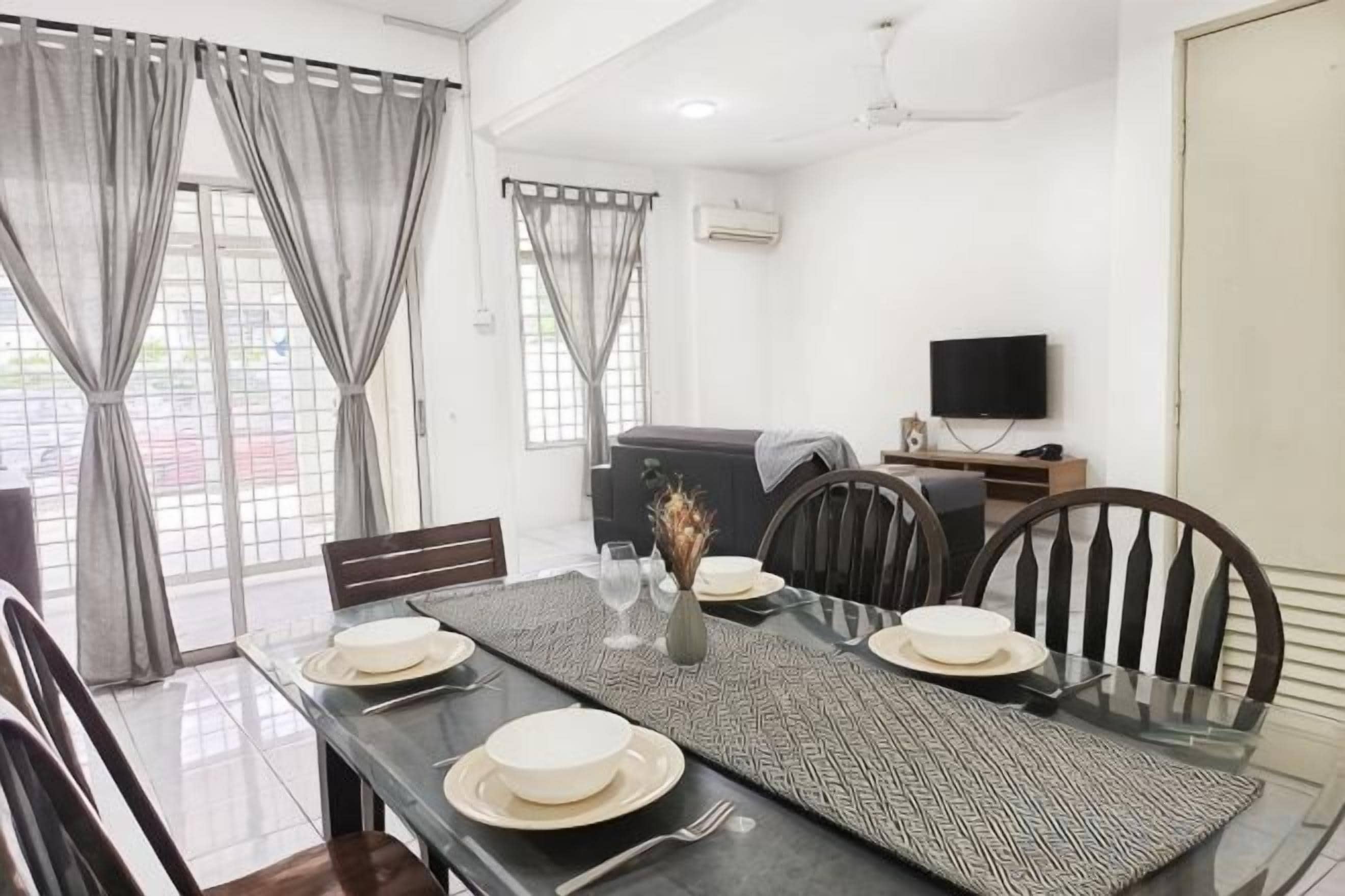 Uma sala de jantar com mesa posta para quatro pessoas, cadeiras, televisão e um sofá ao fundo.