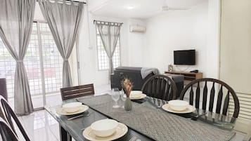 Uma sala de jantar com mesa posta para quatro pessoas, cadeiras, televisão e um sofá ao fundo.
