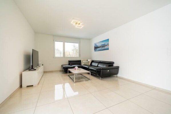 A modern living room with a black leather sofa, a glass coffee table, and a flat-screen TV on a white cabinet. - Peaceful Swiss Retreat Parking + Private Balcony (Safenwil)