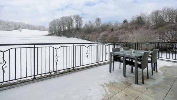 Un patio exterior con mesa y sillas, baranda metálica y vista a un paisaje nevado con árboles.
