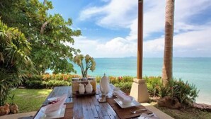 A wooden table set for dining with white dishes, a pitcher, and a vase of flowers, overlooking a clear blue sea. - Baan Chang (Koh Samui)