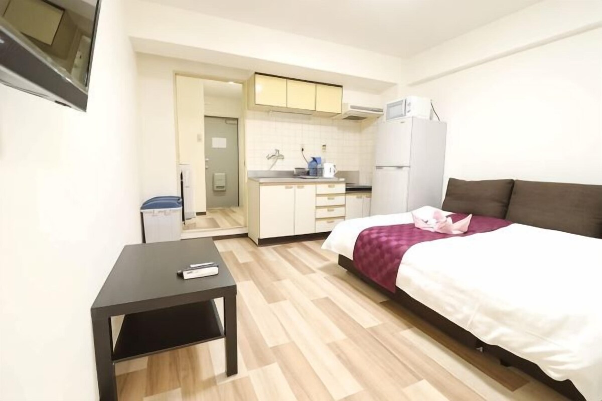 A bedroom with a bed, a small table, a kitchenette, and a bathroom visible through an open door.