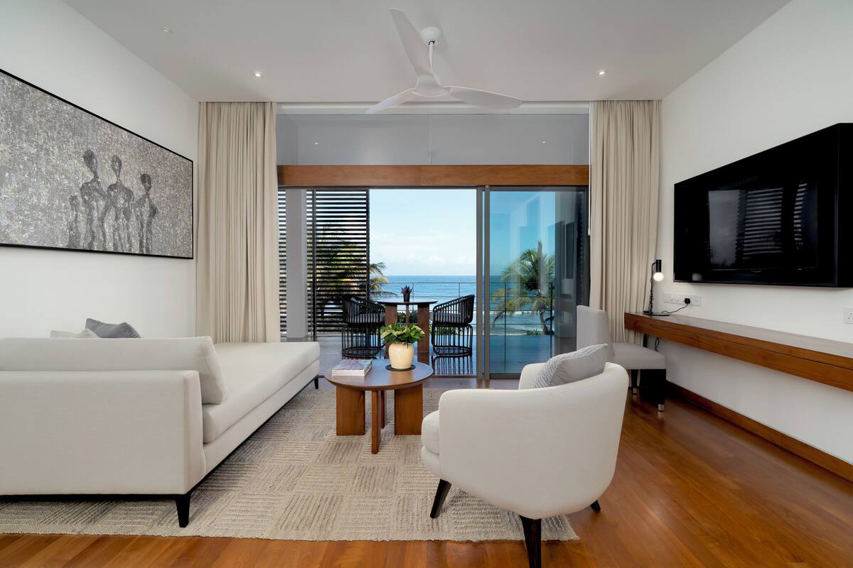 Una sala de estar moderna con un sofá, un sillón y una mesa de centro con una planta. Hay un televisor grande en la pared y un balcón con vistas al océano.