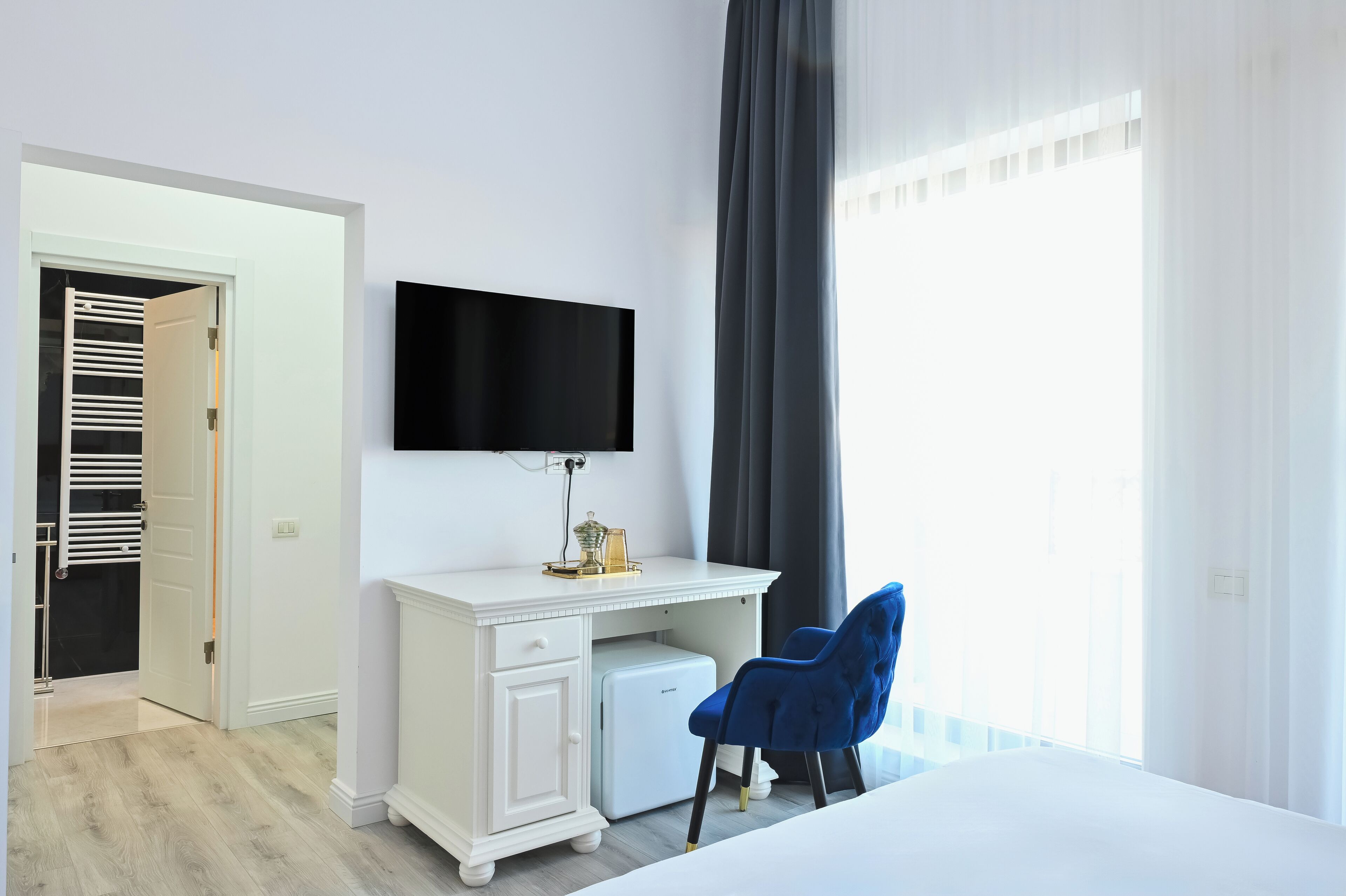 A hotel room with a white desk, a flat-screen TV, a blue chair, and a large window with curtains.