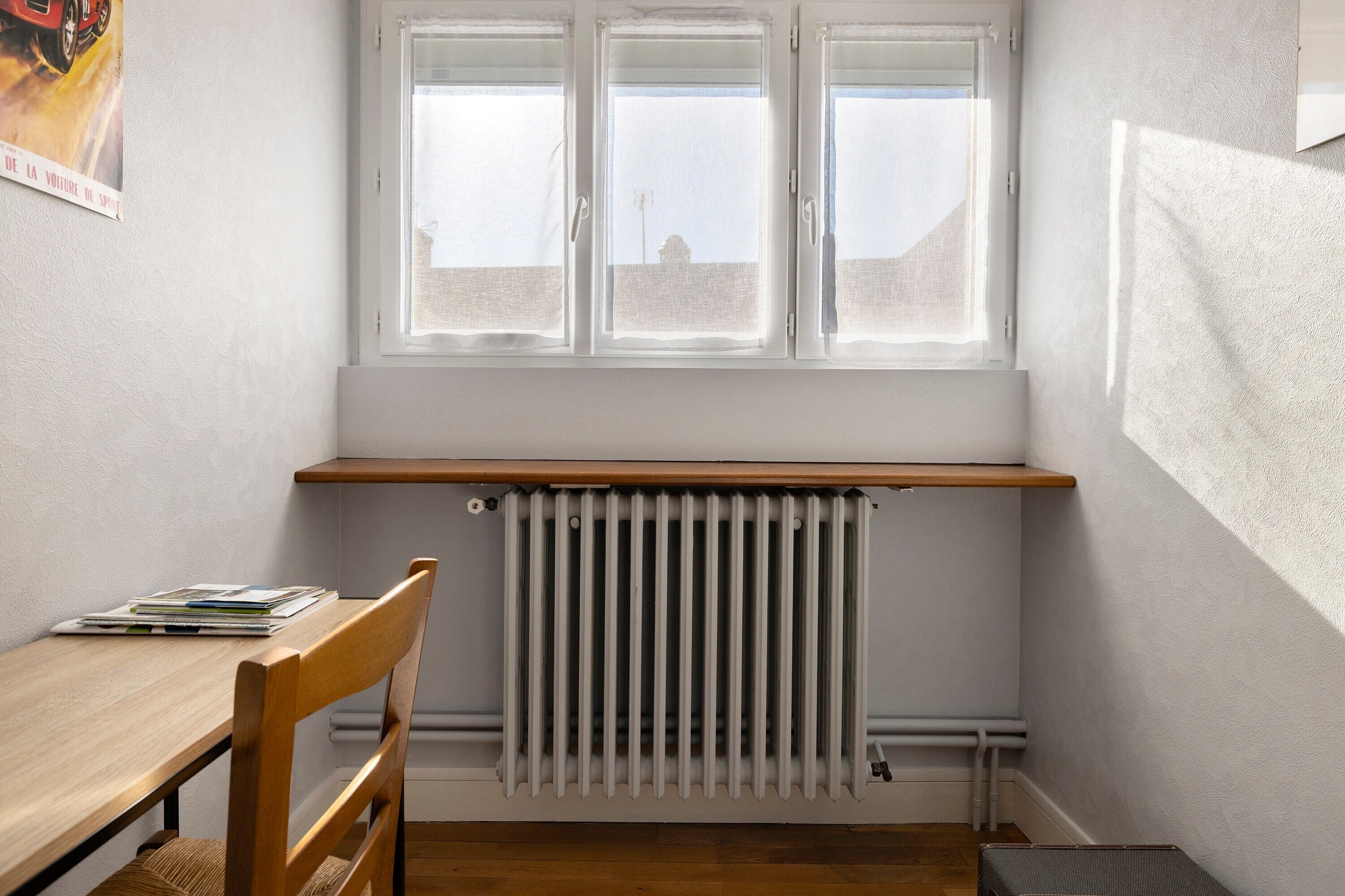 Ein Eckzimmer mit Heizkörper, einem Holzstuhl, einem Tisch mit Büchern und einem Fenster mit Jalousien.