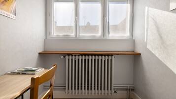 Ein Eckzimmer mit Heizkörper, einem Holzstuhl, einem Tisch mit Büchern und einem Fenster mit Jalousien.