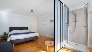 Een moderne slaapkamer met een bed, nachtkastje, klein krukje, douchecabine met glazen deur en een houten vloer.