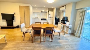 Una cocina moderna con armarios blancos, una mesa de comedor con sillas, un televisor y un balcón con cortinas.