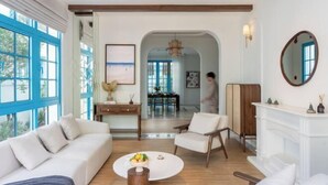 A spacious living room with a white sofa, a coffee table with fruit, a fireplace, and large windows with blue frames. - domdomHOUSE Villa - 1min to beach (Ha Long)