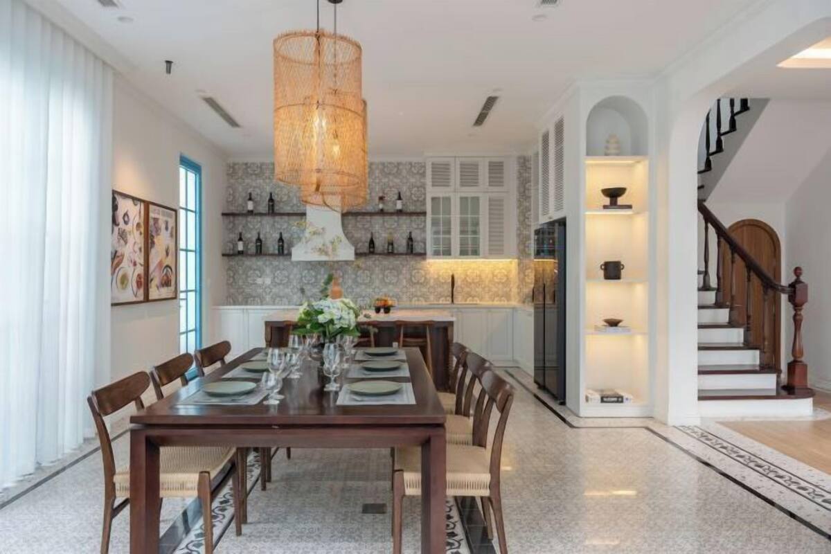 A dining room with a long table set for six, a chandelier, and a kitchen area with shelves.