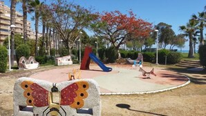 A playground with a slide and climbing structures, surrounded by palm trees and a butterfly mosaic sculpture. - Acv - Aguamarina-2nd line 5th north (Oropesa del Mar)