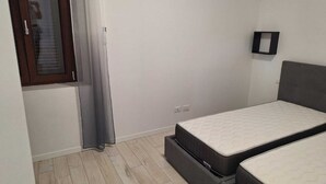 A bedroom with two beds, a window with blinds, a door, and a light switch on the wall. - Starlight Apartment (Garbagnate Milanese)