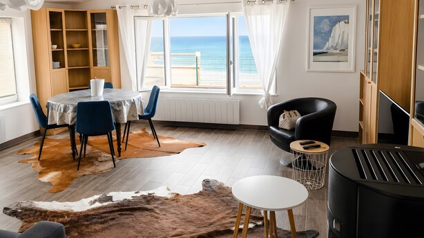 A room with a dining table, chairs, a sofa, a small round table, a heater, a fireplace, a view of the sea, and a bookshelf.