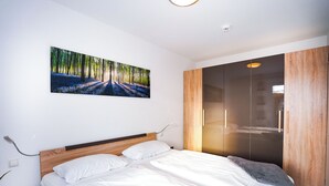 Une chambre moderne avec un grand lit, une tête de lit en bois, un tableau représentant une forêt et une armoire avec un miroir.