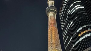 A tall, illuminated tower with a lattice structure and a modern building with vertical lines and lights. - YMK Oshiage 7B (Tokyo)