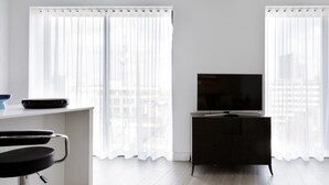 A modern living room with a flat-screen TV on a dark wooden cabinet, a white dining table with a black chair, and sheer white curtains on the windows.