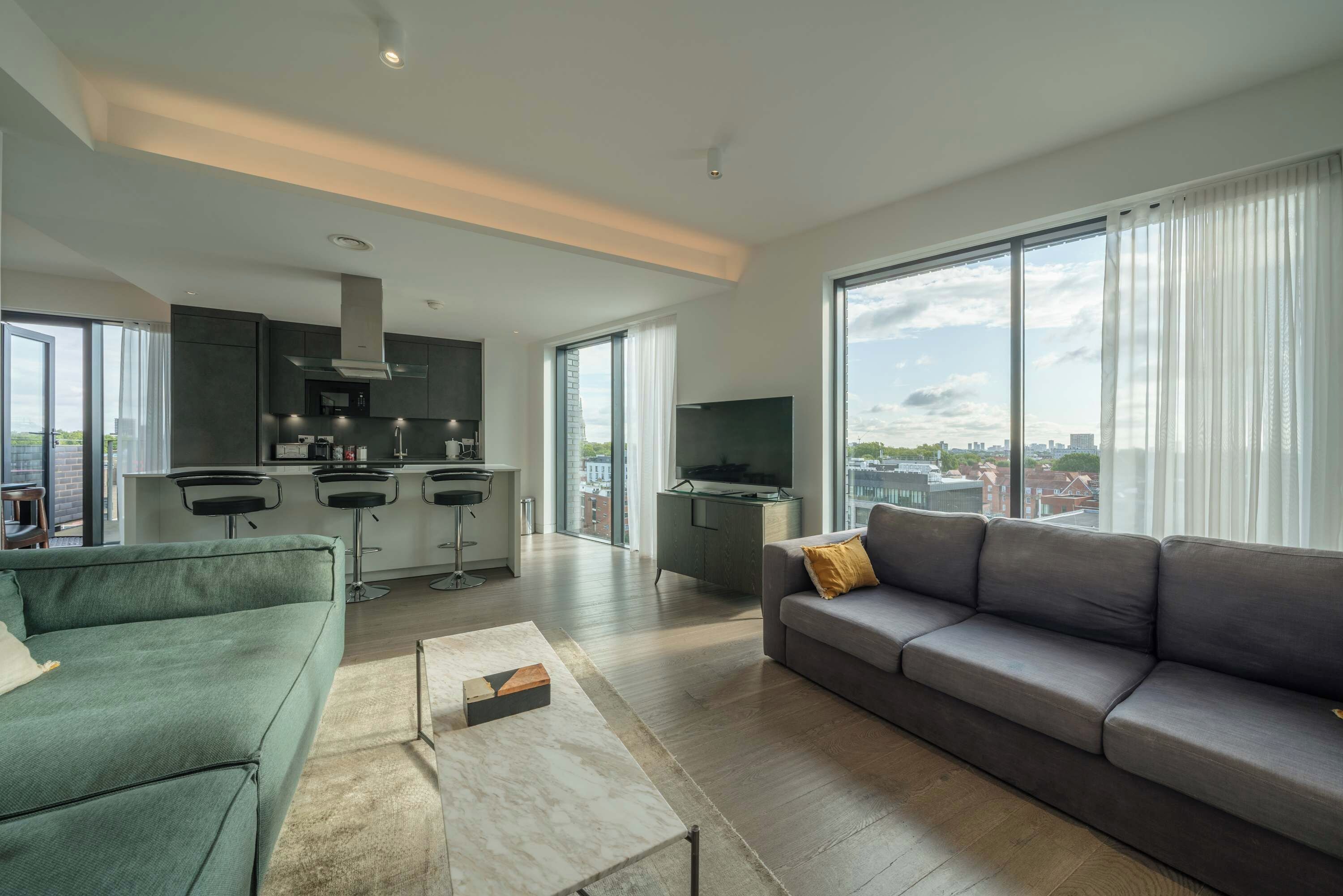Un salon moderne avec un canapé gris, un fauteuil vert et une table basse. Il y a également un coin cuisine avec un bar et un téléviseur. De grandes fenêtres offrent une vue sur la ville.
