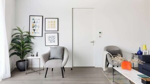 A modern living room with a grey armchair, a small white table, and a potted plant. There are framed artworks on the wall and a door to the right.